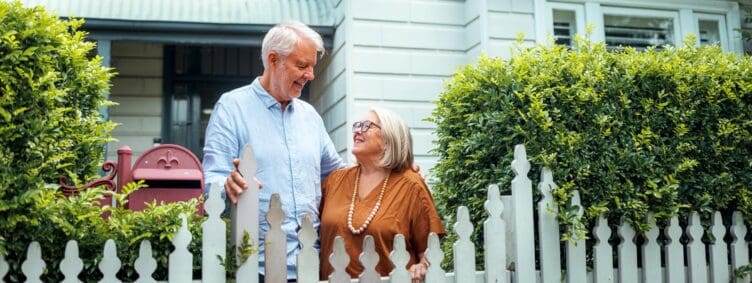 happy-senior-couple-in-front-of-house-with-white-picket-fence
